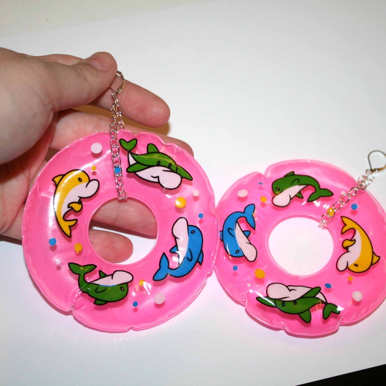 Puede incluir: Un par de pendientes de aro inflables de color rosa con un dise&ntilde;o de delfines y peces. Los aros est&aacute;n unidos a cadenas plateadas con ganchos.