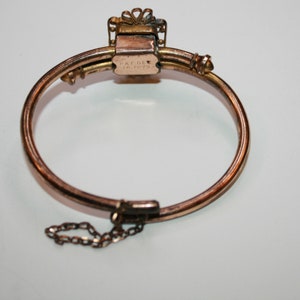 May include: A vintage gold-toned bracelet with a hinged design. The bracelet features a rectangular clasp with the text "PAT. DEC. 16. 1879". A small chain is attached for added security. The bracelet has a decorative top.