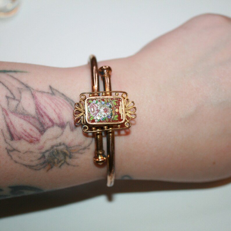 May include: Gold-toned hinged bangle bracelet with a rectangular centerpiece. The centerpiece features a mosaic floral design in shades of red, pink, white, and green. The bracelet is a classic piece of jewelry.