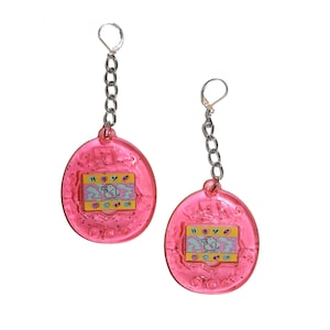May include: A pair of pink plastic earrings shaped like a Tamagotchi, a popular 90s virtual pet toy. Each earring features a silver chain and a clear plastic charm with a colorful pixelated screen and buttons.