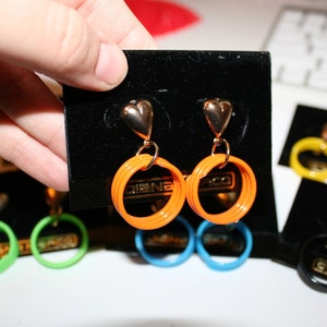 May include: A pair of orange hoop earrings with gold heart-shaped clasps. The earrings are displayed on a black velvet background.