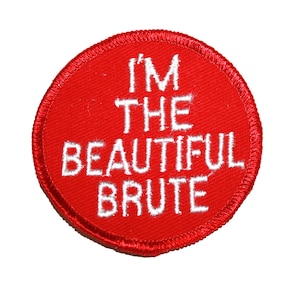 May include: A round, red patch with white embroidered text that reads "I'M THE BEAUTIFUL BRUTE". The patch has a stitched border and is set against a white background.