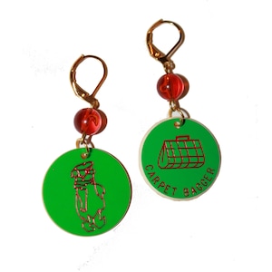 May include: A pair of green and red earrings with a gold hook. One earring features a red cartoon image of a dog wearing a hat and the other earring features a red cartoon image of a suitcase with the text "CARPET BAGGER".