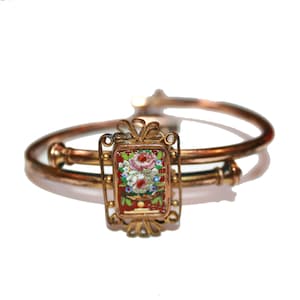 May include: A gold-toned bracelet with a rectangular centerpiece featuring a floral mosaic design. The mosaic showcases pink, white, and blue flowers against a red background. The bracelet has a hinged design.
