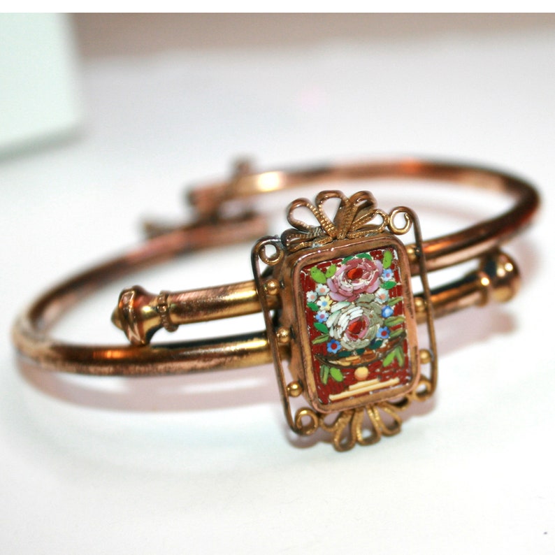 May include: A vintage gold-toned bracelet with a rectangular centerpiece. The centerpiece features a mosaic floral design with red, pink, and white flowers on a red background. The bracelet has a hinged design.