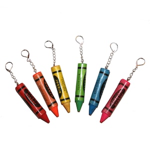 May include: Six colorful crayon-shaped keychains with silver chains. Each keychain has the word "crayon" and "eraser" printed on it. The crayons are red, orange, yellow, green, blue, and pink.