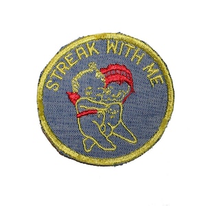 May include: A circular patch with the embroidered text "STREAK WITH ME" in yellow. The patch features a blue background with a yellow border. The image in the center depicts two figures embracing, with red and yellow accents.