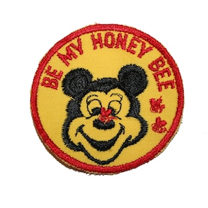 May include: Embroidered patch with a yellow background, a red border, and the text "BE MY HONEY BEE". The patch features a cartoon bee with a smiling face and black ears. Two red flower designs are on the right side.