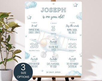 Puede incluir: Un póster de hitos para bebés personalizado con el nombre "JOSEPH" y el texto "¡tiene un año!" El póster incluye detalles como la altura (66 cm), el peso (10 kg), las cosas favoritas y el libro favorito. El diseño presenta un avión azul y estrellas.