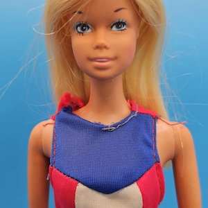 May include: A vintage doll with long blonde hair and blue eyes. The doll wears a red, white, and blue swimsuit. The swimsuit has a blue top, a white center panel, and red sides. The doll is a classic toy.