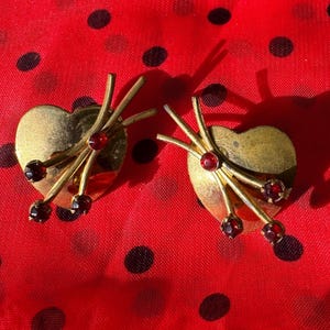 May include: A pair of gold-toned heart-shaped earrings featuring three radiating metal stems, each tipped with a small, round, dark red gemstone. The earrings have a polished metallic finish and a symmetrical design with a smooth, curved heart base.