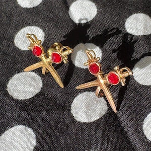 May include: A pair of gold-colored earrings, each featuring a sword design with a red gemstone at the center. The earrings are set against a black and white polka dot background, creating a striking contrast.