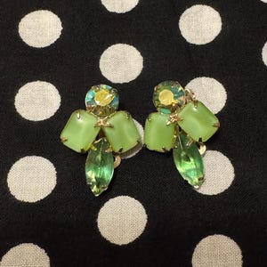 May include: A pair of clip-on earrings featuring a cluster of light green rectangular stones, a single marquise-cut translucent green stone at the base, and a round iridescent yellow stone at the top, all set in a gold-toned metal frame.