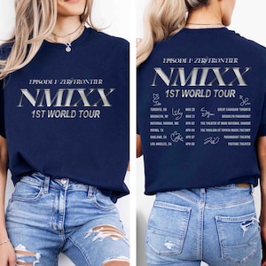 May include: Navy blue t-shirt with the text "EPISODE 1: ZERO FRONTIER" and "NMIXX 1ST WORLD TOUR" in silver lettering. The back of the shirt features a tour schedule with city names and dates. The shirt is worn with blue jeans.