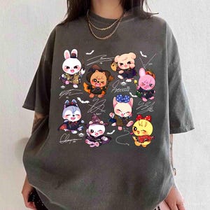 May include: Dark grey t-shirt featuring a colorful cartoon graphic of nine animal characters in Halloween costumes. Each character has unique outfits and accessories. The design includes bats and signatures.