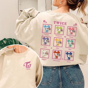 May include: Beige sweatshirt featuring the text "TWICE" and a graphic of nine cartoon characters in postage stamp designs. The left sleeve has the "Twice" logo. The sweatshirt is made of a soft material.