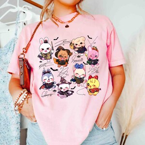 May include: Pink t-shirt featuring a colorful graphic of cartoon animals in costumes, with signatures and bat illustrations. The shirt is a light pink color and has a relaxed fit.