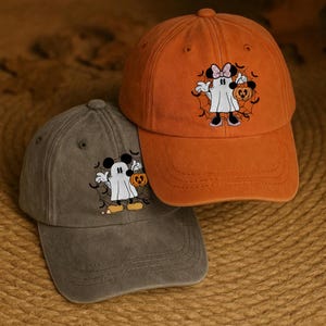 Puede incluir: Dos gorras de béisbol, una naranja y otra gris, con un diseño temático de Halloween. Las gorras tienen un fantasma de dibujos animados bordado sosteniendo una calabaza. La gorra naranja tiene un lazo rosa en el fantasma. Las gorras están sobre una superficie tejida.