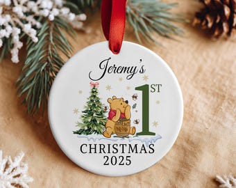 Op de afbeelding: Ronde witte kerstversiering met een rood lint. De versiering heeft een cartoonbeer met een honingpot, een kerstboom en de tekst "Jeremy's 1st Christmas 2025".
