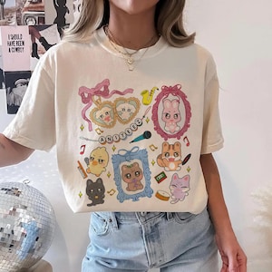 May include: Cream-colored t-shirt featuring a colorful graphic design. The design includes framed cartoon animals, musical instruments, and the word "ANITEEZ". The shirt has short sleeves and a crew neck.