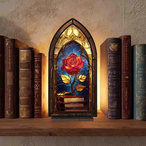 May include: A stained glass-style lamp featuring a red rose with gold leaves and stem, set against a blue starry background. The lamp is in a gothic arch frame, resting on a stack of books. The lamp is on a wooden shelf with antique books.