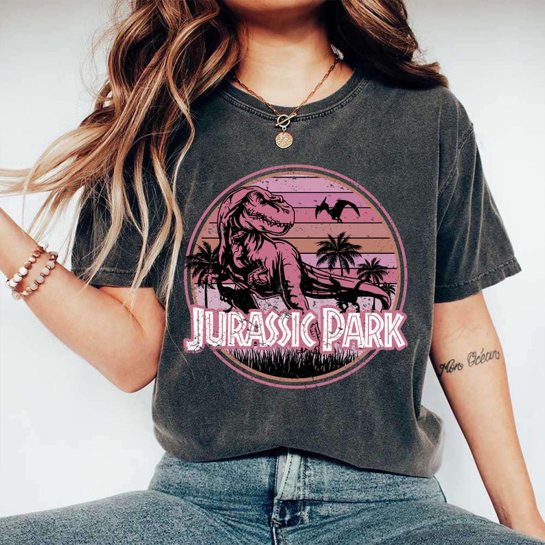 Pu&ograve; includere: T-shirt grigio scuro con una grafica rosa e viola di un Tyrannosaurus Rex all'interno di un design circolare. La grafica include palme, un tramonto e il testo "JURASSIC PARK" in rosa.