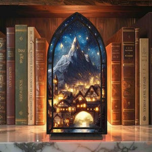 Op de afbeelding: Een decoratief glas-in-loodpaneel met een bergdorpscène onder een sterrenhemel. Het paneel is zwart omlijst en staat op een plank met antieke boeken. De boeken hebben bruine en groene covers.