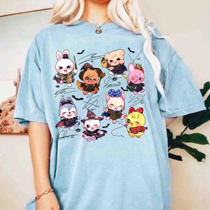 May include: Light blue t-shirt featuring a graphic of cartoon animals in Halloween costumes. The design includes a bunny with a wand, a bear in a cape, and a cat in a witch hat. The shirt also has signatures and bat illustrations.