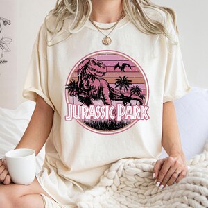 Pu&ograve; includere: T-shirt color crema con grafica circolare raffigurante un tramonto rosa e arancione. La grafica include un Tyrannosaurus Rex, palme e la scritta "JURASSIC PARK" in rosa. La maglietta &egrave; di stile casual.