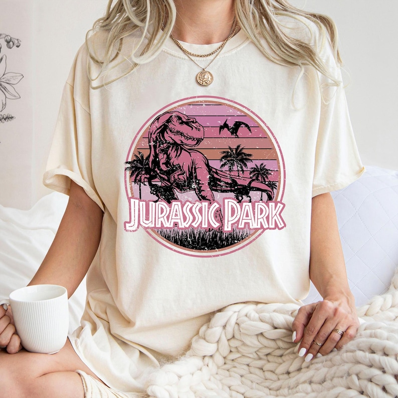 Pu&ograve; includere: T-shirt color crema con grafica circolare raffigurante un tramonto rosa e arancione. La grafica include un Tyrannosaurus Rex, palme e la scritta "JURASSIC PARK" in rosa. La maglietta &egrave; di stile casual.