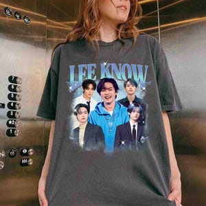 May include: Dark gray t-shirt featuring a graphic of a group of young men with the text "LEE KNOW" in a light blue, vintage-style font. The image has a faded, retro aesthetic.