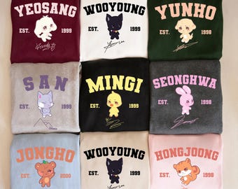 May include: Nine sweatshirts in different colours, each with a name in bold lettering, a cartoon character, and the year 1998, 1999, or 2000. The sweatshirts include names such as Yeosang, Wooyoung, Yunho, San, Mingi, Seonghwa, Jongho, and Hongjoong.