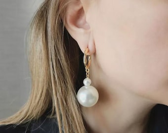 May include: Gold hoop earrings with a large, white, pearl-like bead suspended from each hoop. A smaller pearl-like bead is attached above the larger bead. The earrings are shown against a blurred background.