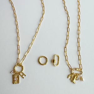 May include: Gold-toned necklaces with various charms, including a rectangular pendant with a starburst design, a letter "H", and a small crystal. Additional gold accessories include a round ring and a carabiner-style clasp.