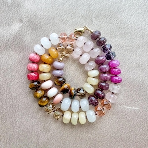 Op de afbeelding: Een cirkelvormige armband met een verscheidenheid aan gladde, ronde edelsteenkralen in tinten wit, geel, roze, bruin, paars en magenta, afgewisseld met gefacetteerde heldere en amberkleurige glaskralen, afgewerkt met een goudkleurige metalen sluiting.