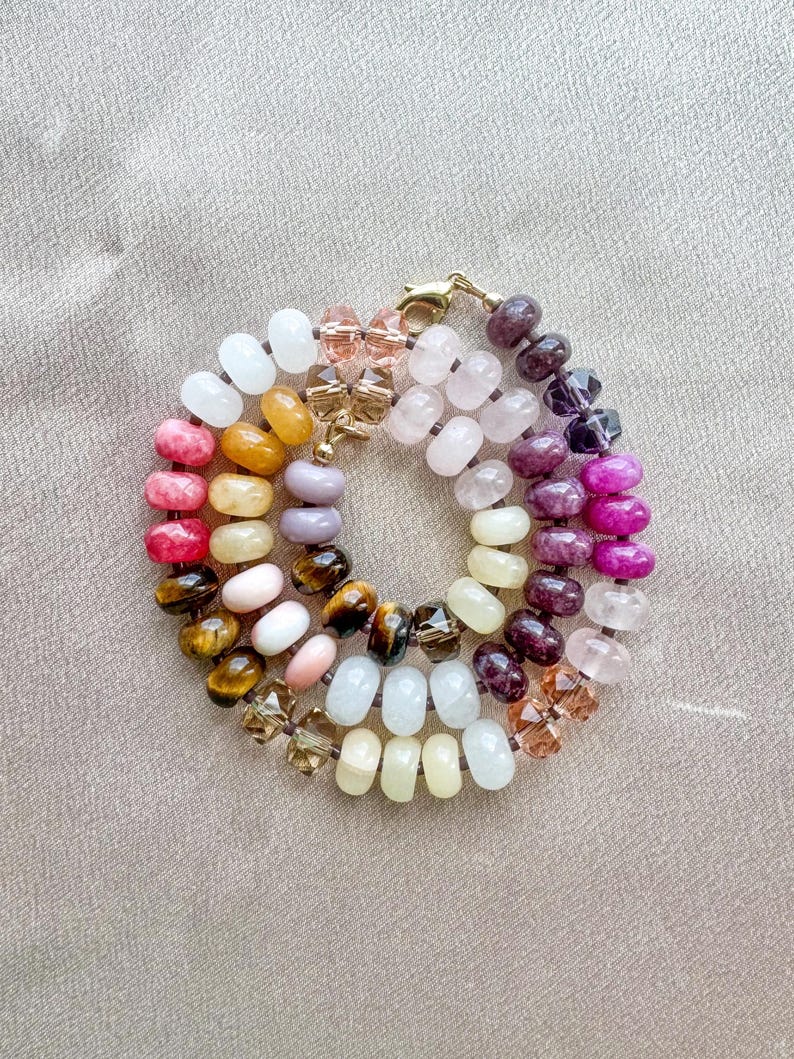 Op de afbeelding: Een cirkelvormige armband met een verscheidenheid aan gladde, ronde edelsteenkralen in tinten wit, geel, roze, bruin, paars en magenta, afgewisseld met gefacetteerde heldere en amberkleurige glaskralen, afgewerkt met een goudkleurige metalen sluiting.