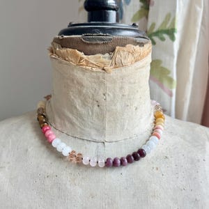 Op de afbeelding: Een kralenketting met een verscheidenheid aan ronde, gladde en gefacetteerde kralen in tinten roze, wit, donkerrood, bruin en geel. De kralen zijn in een reeks op een dun koord geregen, wat zorgt voor een kleurrijk en gestructureerd ontwerp.