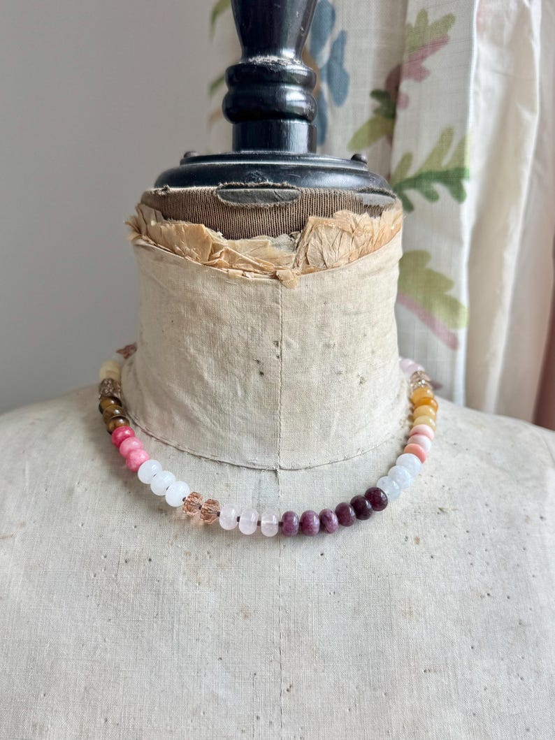Op de afbeelding: Een kralenketting met een verscheidenheid aan ronde, gladde en gefacetteerde kralen in tinten roze, wit, donkerrood, bruin en geel. De kralen zijn in een reeks op een dun koord geregen, wat zorgt voor een kleurrijk en gestructureerd ontwerp.
