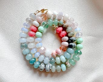 Op de afbeelding: Een meerstrengs ketting met een verscheidenheid aan gladde, ronde en gefacetteerde edelsteenkralen in tinten roze, groen, blauw, wit en grijs. De kralen zijn aan elkaar geregen met een goudkleurige metalen karabijnsluiting aan de bovenzijde.