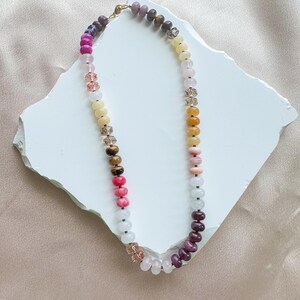 Op de afbeelding: Een kralenketting met een verscheidenheid aan ronde, gladde en gefacetteerde kralen in tinten roze, geel, wit, bruin en paars. De ketting is geregen aan een dun koord en bevat een goudkleurige metalen sluiting aan de bovenkant om de uiteinden vast te maken.