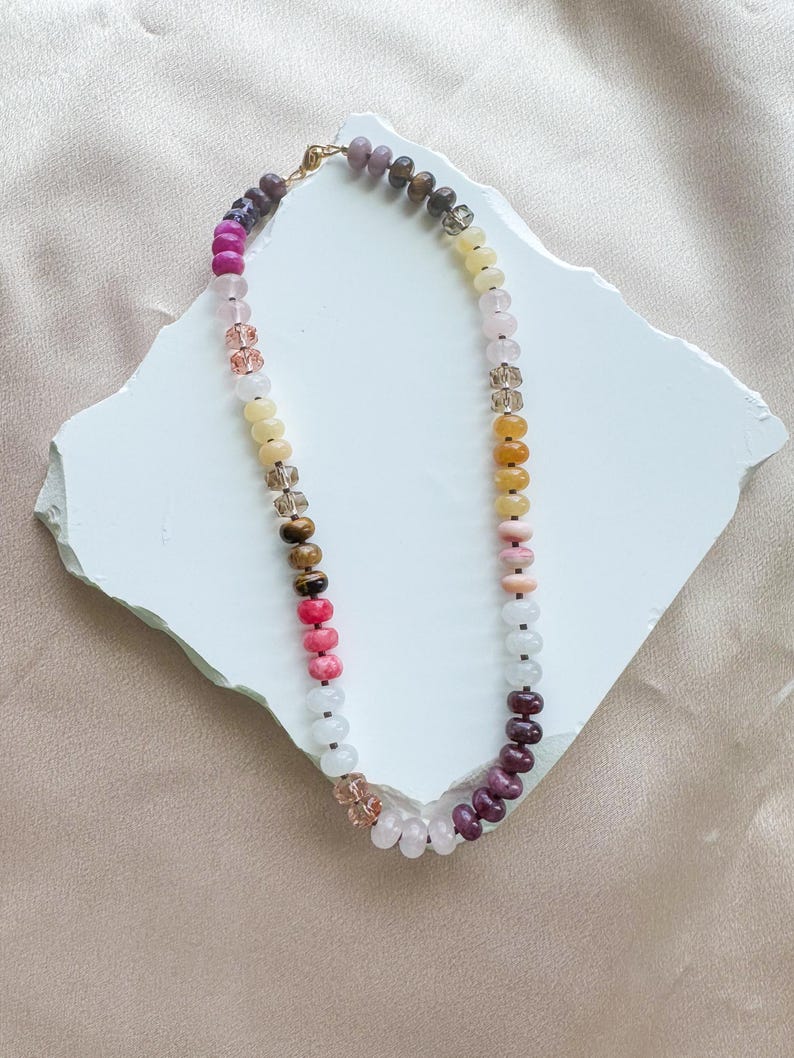 Op de afbeelding: Een kralenketting met een verscheidenheid aan ronde, gladde en gefacetteerde kralen in tinten roze, geel, wit, bruin en paars. De ketting is geregen aan een dun koord en bevat een goudkleurige metalen sluiting aan de bovenkant om de uiteinden vast te maken.