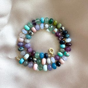 K&ouml;nnte beinhalten: Eine Halskette bestehend aus bunten, runden, facettierten Edelsteinperlen in den Farben Rosa, Blau, Gr&uuml;n, Violett, Schwarz und Wei&szlig;. Die Perlen sind aufgereiht und mit einem goldfarbenen Karabinerverschluss sowie einem Biegering befestigt.