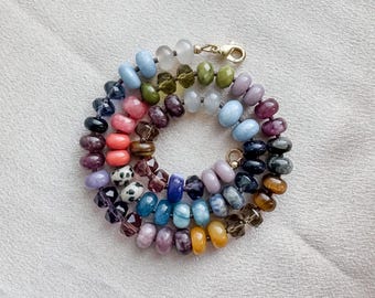 Könnte beinhalten: Eine mehrfarbige Perlenkette mit einer Vielzahl von glatten, runden und facettierten Edelsteinperlen in Blau-, Rosa-, Lila-, Grün-, Gelb- und Schwarztönen, die in einem Spiralmuster angeordnet und mit einem goldfarbenen Karabinerverschluss versehen ist.