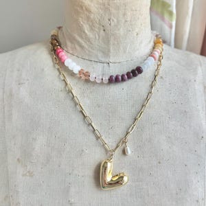 Op de afbeelding: Een gelaagde ketting met een bovenste streng van veelkleurige ronde kralen in roze, witte en donkerrode tinten, en een onderste goudkleurige ketting met een grote, gladde, gepolijste goudkleurige hartvormige hanger en een kleine witte parelbedel.