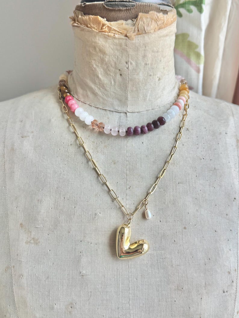 Op de afbeelding: Een gelaagde ketting met een bovenste streng van veelkleurige ronde kralen in roze, witte en donkerrode tinten, en een onderste goudkleurige ketting met een grote, gladde, gepolijste goudkleurige hartvormige hanger en een kleine witte parelbedel.