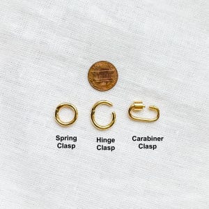 Op de afbeelding: Drie goudkleurige sieradenonderdelen horizontaal gerangschikt onder een koperen munt. De items zijn gelabeld als een ronde veerring, een ronde scharniersluiting en een ovale karabijnsluiting, elk met een functioneel openingsmechanisme.