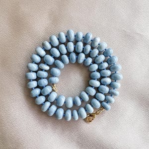 May include: A necklace composed of numerous smooth, round, light blue beads strung together in a circular pattern. The beads feature subtle variations in color and texture. The necklace is secured with a gold-toned metal lobster claw clasp.