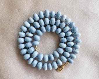 Op de afbeelding: Een ketting bestaande uit talloze gladde, ronde, lichtblauwe kralen die in een cirkelvormig patroon zijn geregen. De kralen vertonen subtiele variaties in kleur en textuur. De ketting is voorzien van een goudkleurige metalen karabijnsluiting.