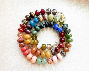 Op de afbeelding: Een cirkelvormige ketting bestaande uit veelkleurige, gefacetteerde en gladde ronde kralen in tinten rood, oranje, geel, groen, blauw en roze. De streng heeft een goudkleurige metalen karabijnsluiting en is in een spiraalvorm gelegd.