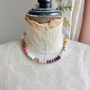 Op de afbeelding: Een kralenketting met een verscheidenheid aan gladde, ronde edelsteenkralen in tinten roze, wit, dieppaars, bruin en doorschijnend amber. De kralen zijn gerangschikt in een doorlopend cirkelvormig patroon rond de nek van een vintage paspop.